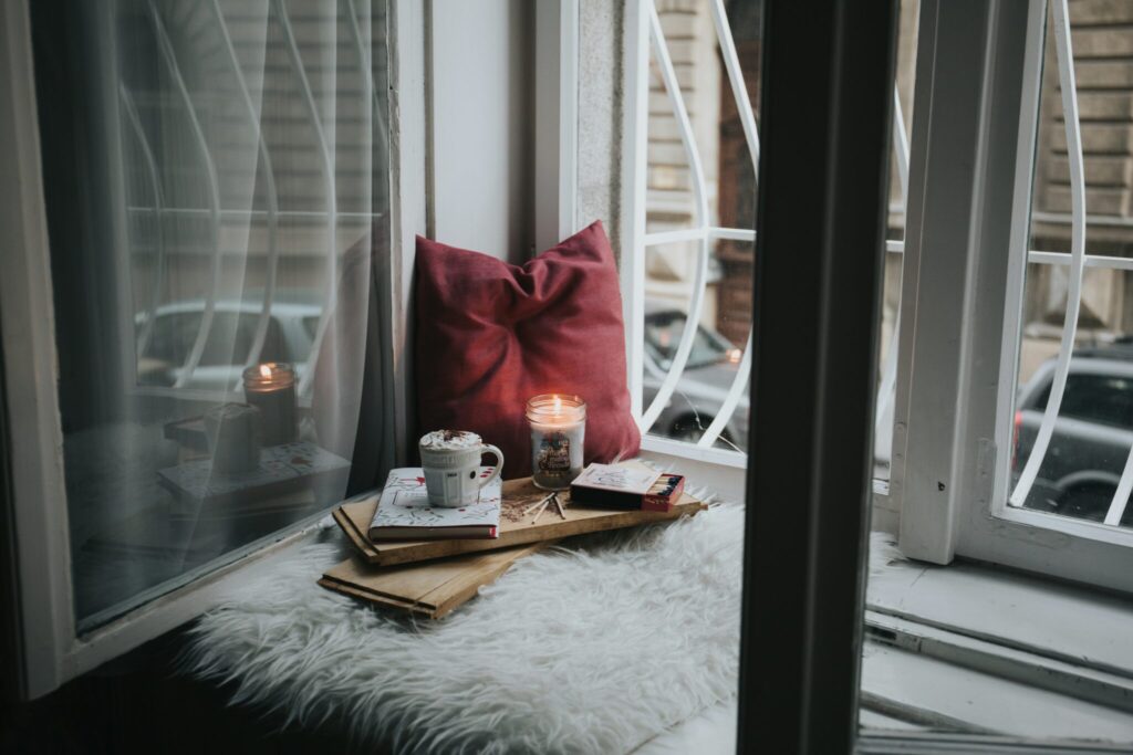 A warm, inviting windowsill setup with a candle, books, a red pillow, and a mug of hot chocolate on a soft white blanket.