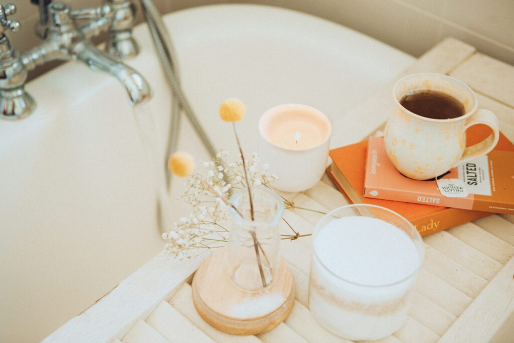 A calming bath setup featuring a lit candle, a mug of tea on stacked books, and a small vase of flowers arranged on a wooden bath tray.